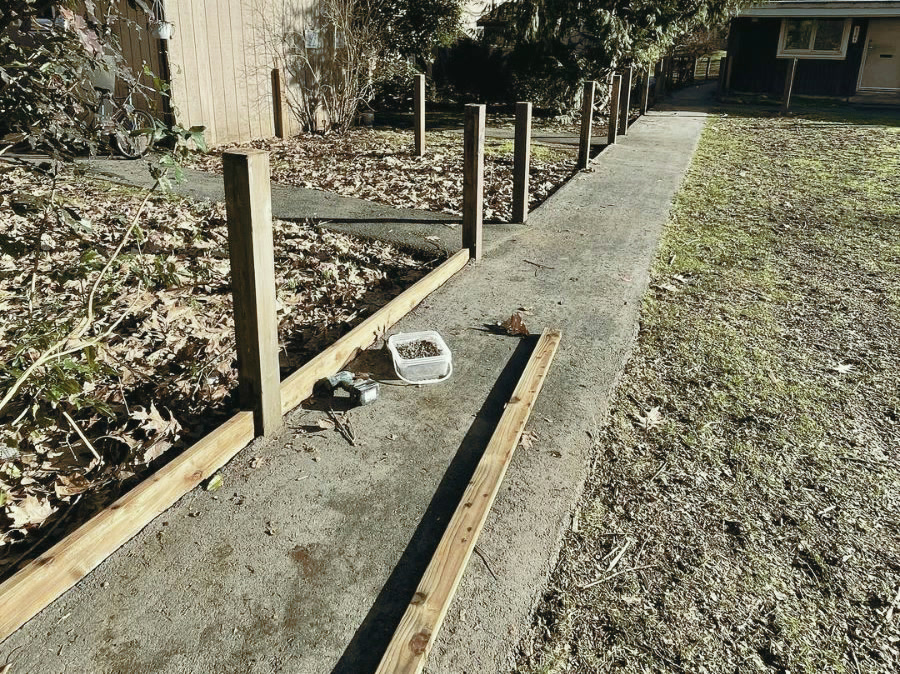 Posts standing along a shared walkway, with pressure-treated rails and kickboards staged on the ground beside them.