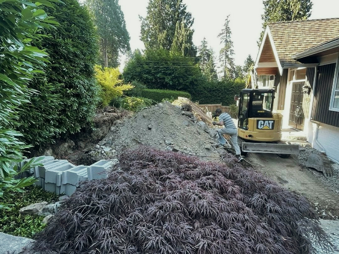 A mini excavator working beside the house, cutting into the slope, with excavated soil stockpiled and retaining wall blocks staged at the edge of the garden.