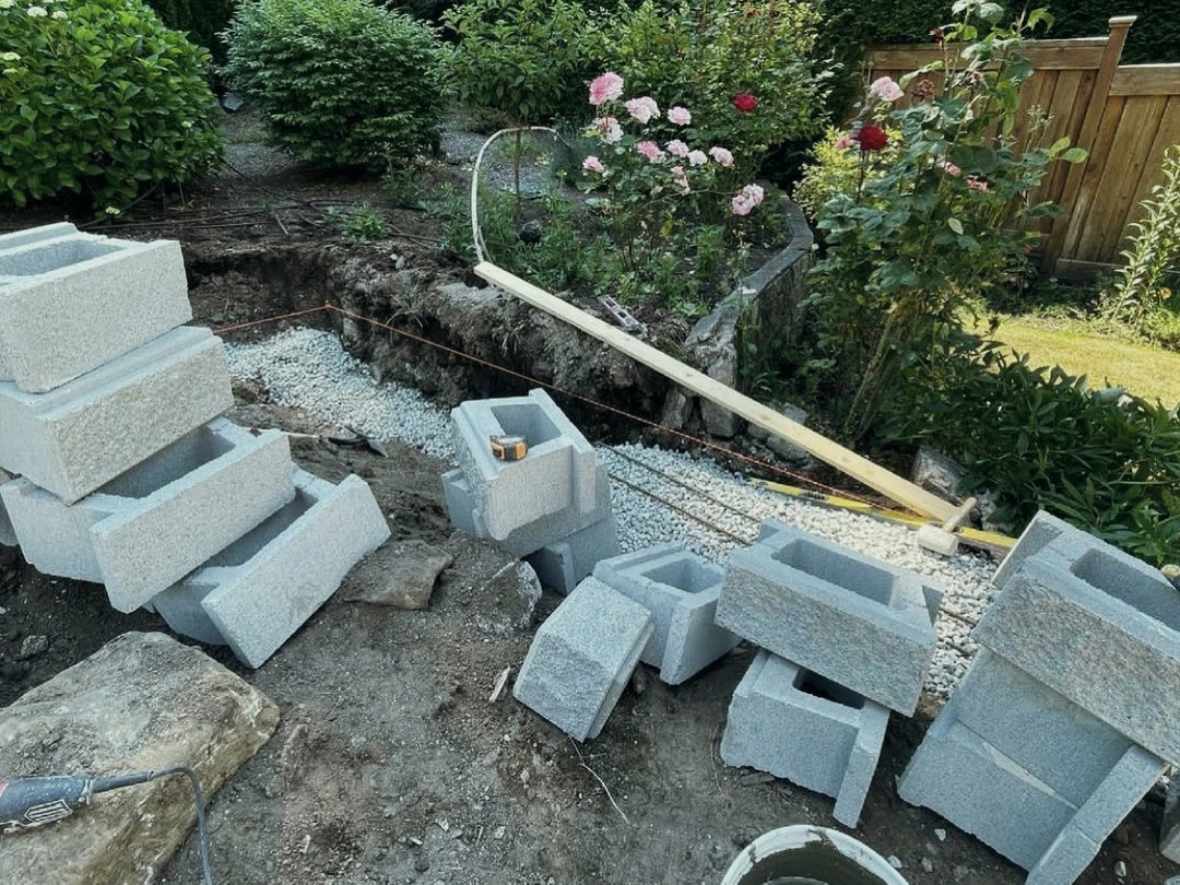 Retaining wall blocks staged beside a cut trench, with a crushed gravel base levelled to a string line ready for the first course.