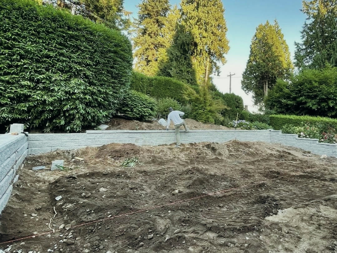 The retaining wall built up along the back of the cut, with a string line run across the excavated pad and soil being backfilled behind the wall.