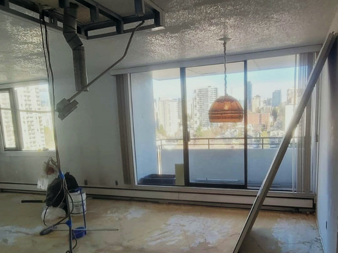 The living area stripped to the concrete slab, the ceiling opened up to steel stud framing and ductwork, and the city visible through the balcony glazing.